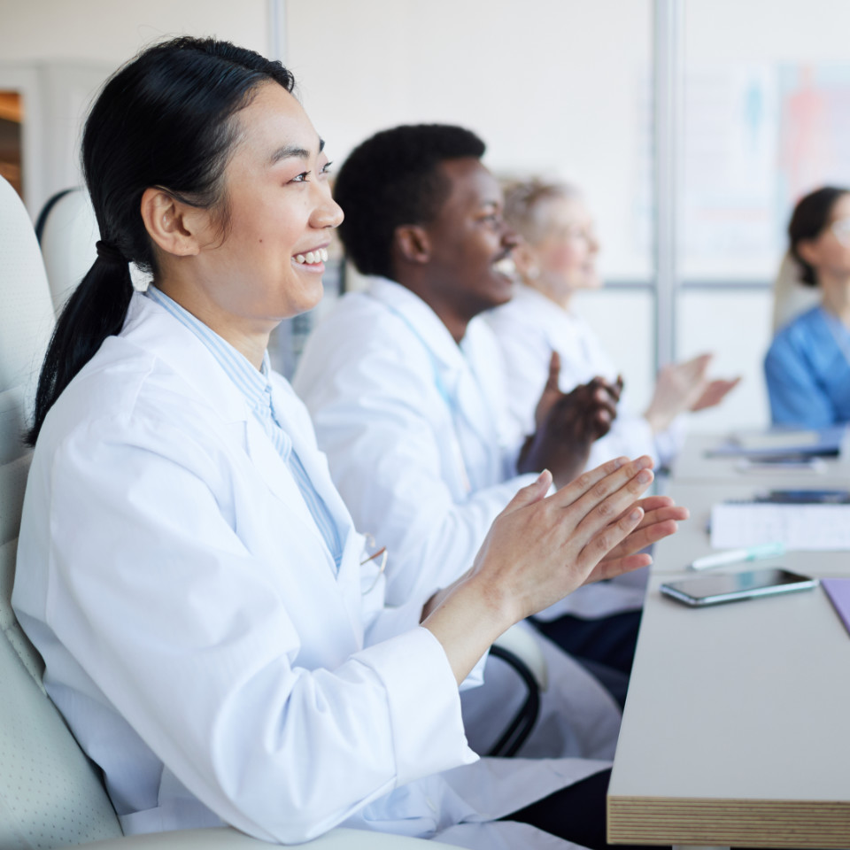 Four medical professionals in white lab coats are seated at a table, smiling and clapping during what appears to be a meeting or presentation.