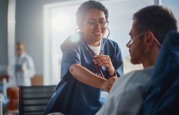 A nurse in scrubs uses a stethoscope to check a male patient’s chest in a hospital room. The patient is sitting up and wearing an oxygen tube.