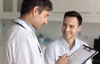 A doctor holds a clipboard and writes while talking to a smiling male patient in a medical office.