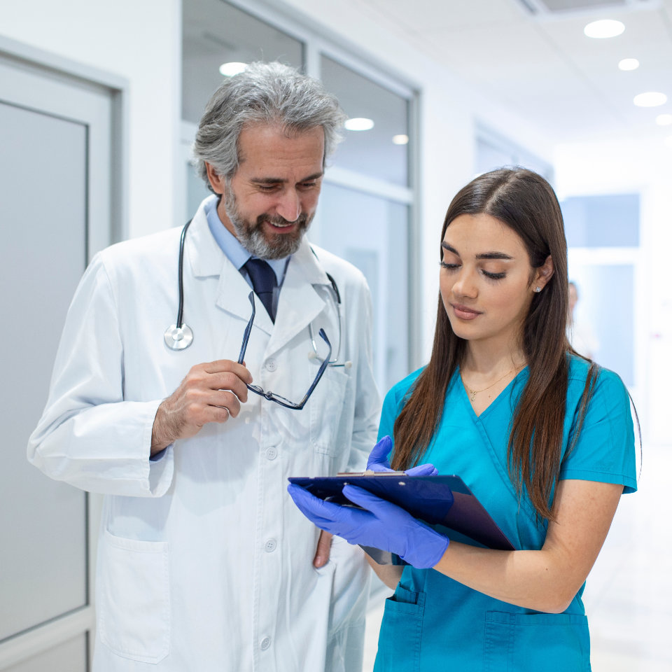 A male doctor and a female nurse in medical uniforms discuss patient information while looking at a clipboard in a hospital hallway.